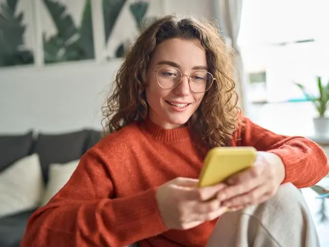A woman in an orange jumper smiling while using a phone with a yellow case.
