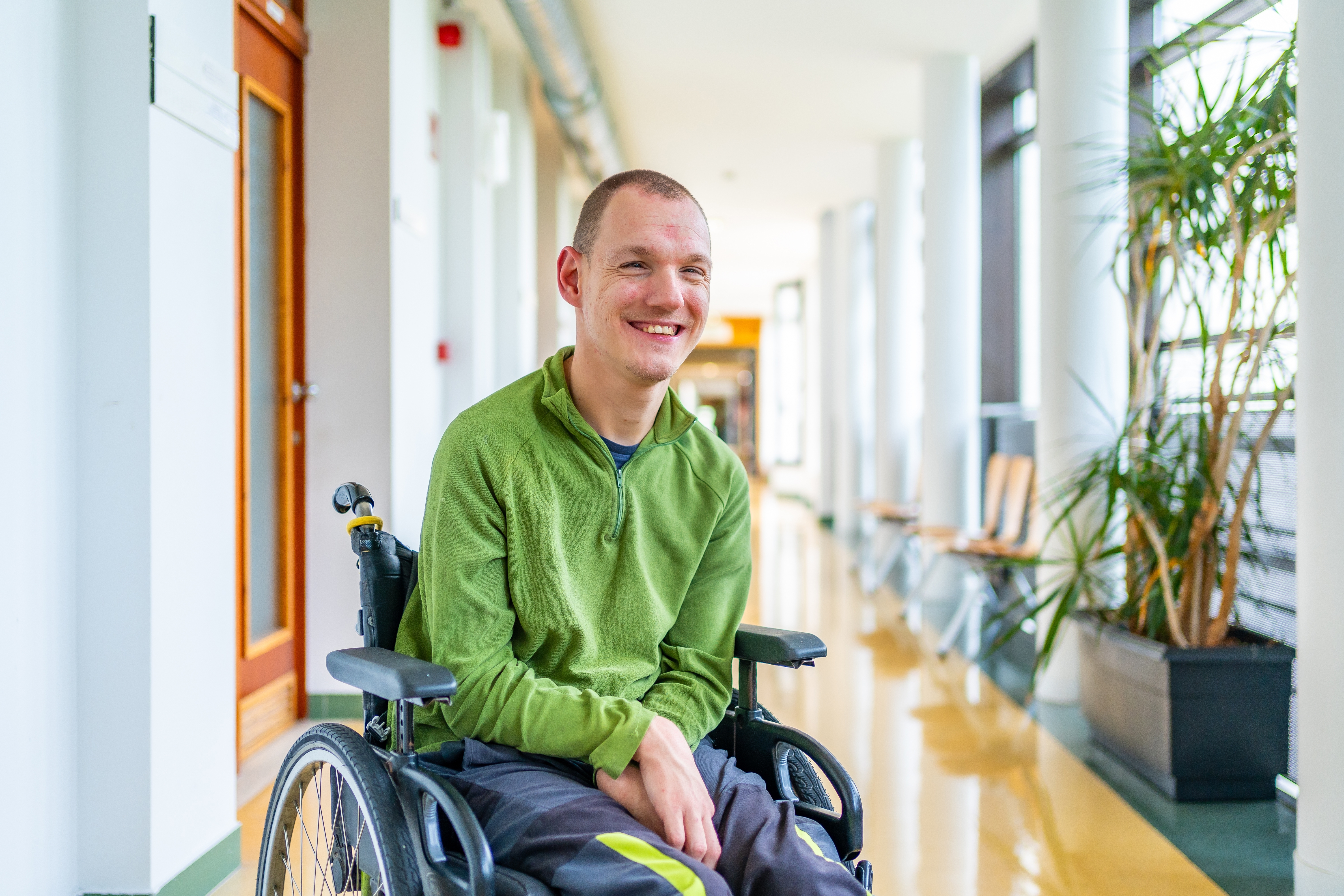 A wheelchair user wearing a green shirt smiling in a corridor of a building