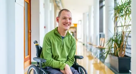 A wheelchair user wearing a green shirt smiling in a corridor of a building