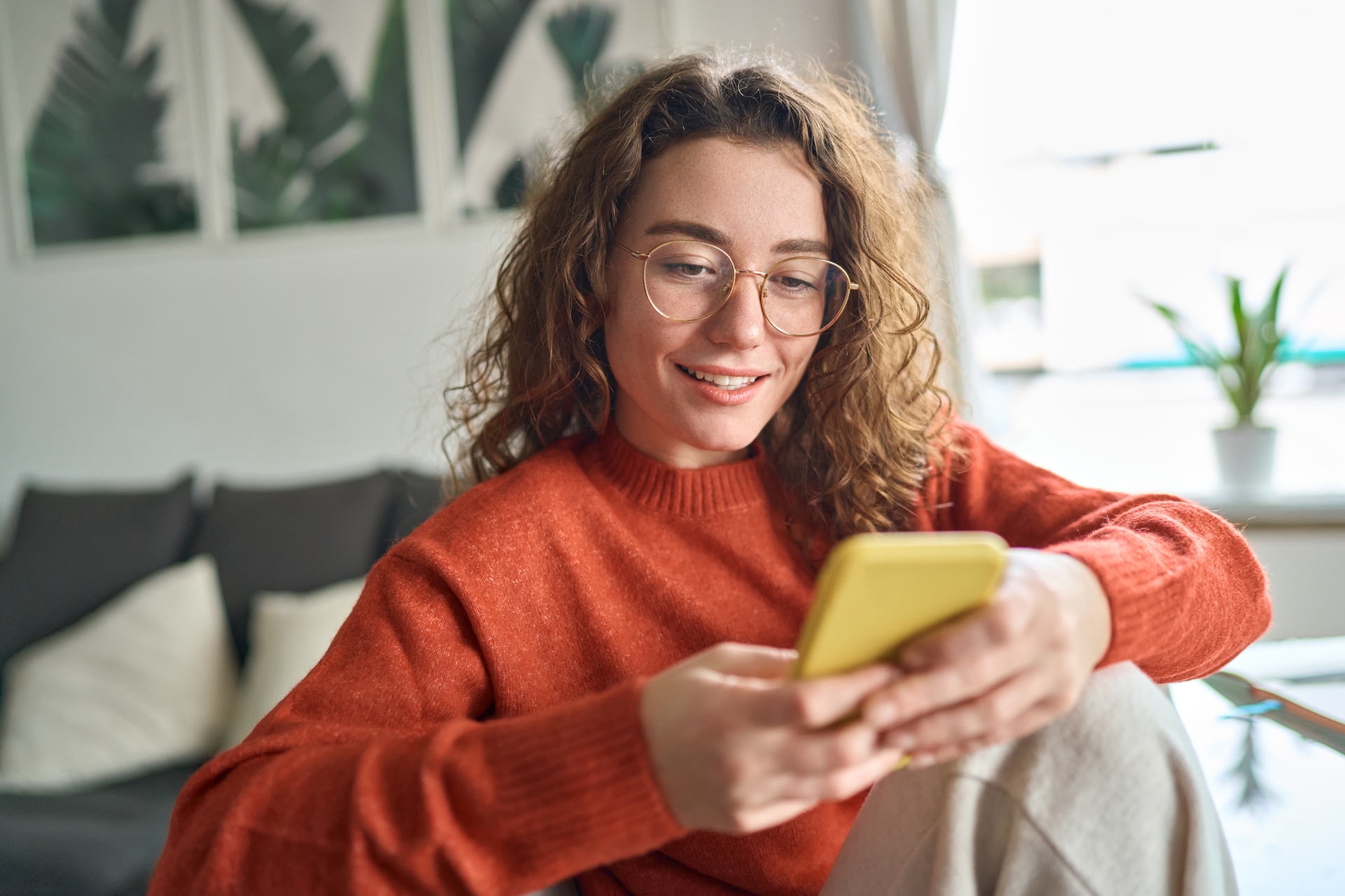 A woman wearing an orange jumper smiling while using a phone with a yellow case
