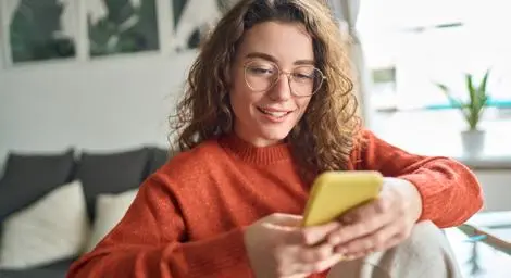 A woman wearing an orange jumper smiling while using a phone with a yellow case